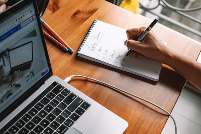 A person writing notes while researching online courses on a laptop in a home setting.